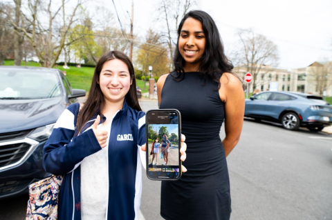 Daphne Garcia, A24, left, and Ayesha Lobo, A24, right, holding an iPhone displaying a photo they took together on their first day at Tufts as first-year students in 2020 while simultaneously recreating the photo on their last day of classes as seniors in 2024.