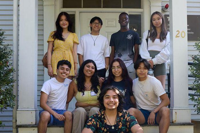 A group of nine young adults smile for a photo while gathered on the front steps of a white building with columns. Five people stand on the top step, three sit on the middle step, and one sits in front. They wear casual summer clothing, and the building entrance is framed by shrubs on either side.
