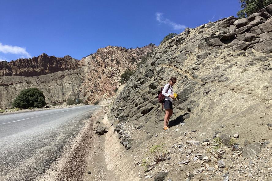 A man with a backpack standing on a rocky terrain by the roadside taking pictures of the rocks