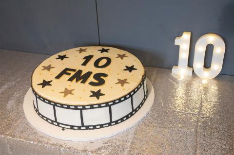 Round white cake celebrating 10 years of FMS, decorated with black and gold stars and film reel detailing, beside illuminated number 10 lights on a silver tablecloth
