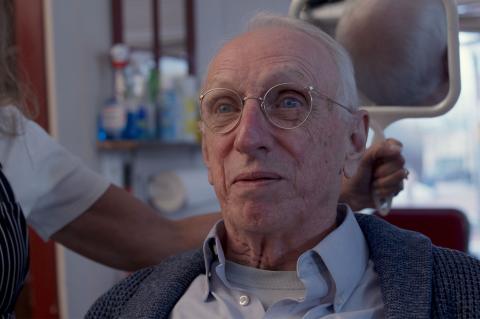 Close-up of an older man wearing round glasses seated in a barber chair while someone adjusts his cape in a barbershop setting