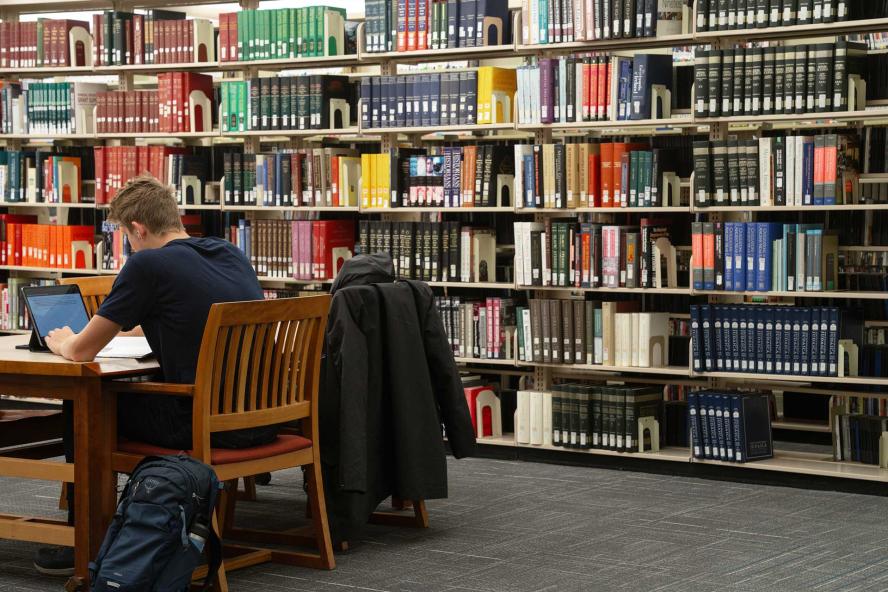 Student using a laptop at a desk alongside rows of books in Tisch Library