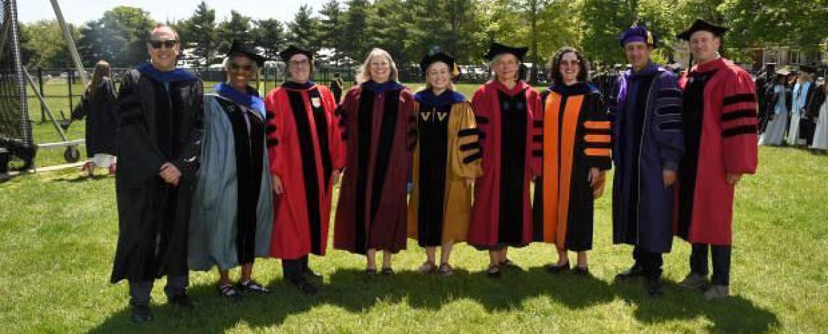 People in academic regalia standing outdoors on a grassy field during a graduation ceremony