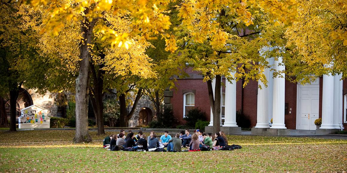 Students, sitting on the ground in a circle, attending a lecture outdoors.