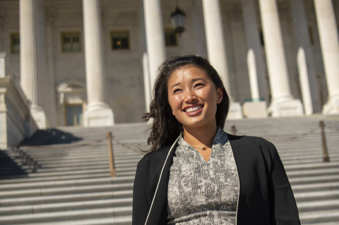 Smiling in dark blazer and gray dress front of white government building with pillars