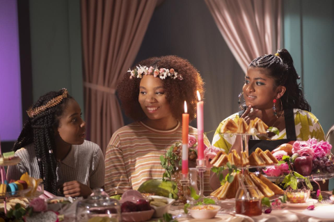 Three young women of color sit behind a table loaded with festive food and candles