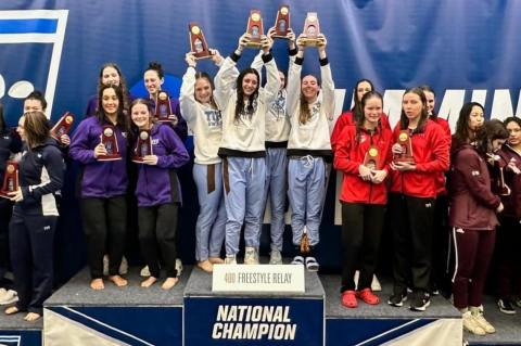 The four women swimmers of the Tufts 400 free relay team hold trophies on the top platform, with other teams to either side of them. Tufts wins three NCAA swimming titles at national competition.