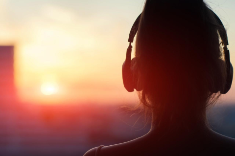 A woman wearing over-the-ear headphones seen in profile from the rear, looking at a setting sun. A music engineering expert explains how to get the best audio quality from streaming music services