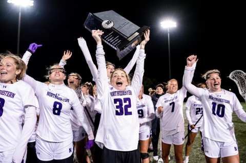 Molly Laliberty, A22, holds a big trophy in the air, surrounded by women’s lacrosse teammates. 