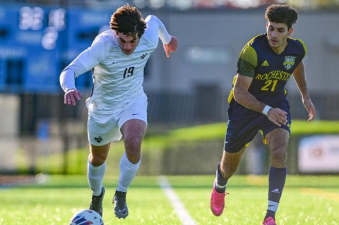 A Tufts soccer player in white kicking a soccer ball ahead of him, while he is pursued by an opposing soccer player. Tufts men’s soccer, women’s soccer, men’s cross country, and individual women runners are in the thick of NCAA postseason competition
