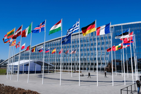 A circle of flagpoles with more than 30 national flags flying in front of a large building that curves toward the ground on the left.