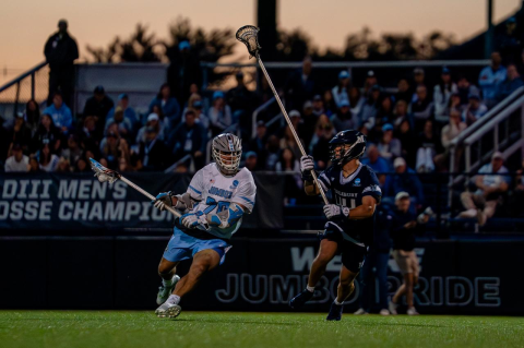 A men’s lacrosse player in a blue and white uniform moves up field against a player in a dark blue uniform.