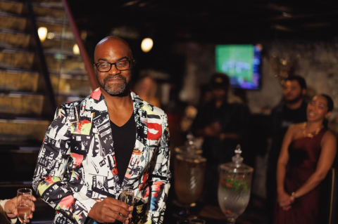 A man stands in a front of a busy room, looking off to the side. He has a vibrant, patterned suit jacket on